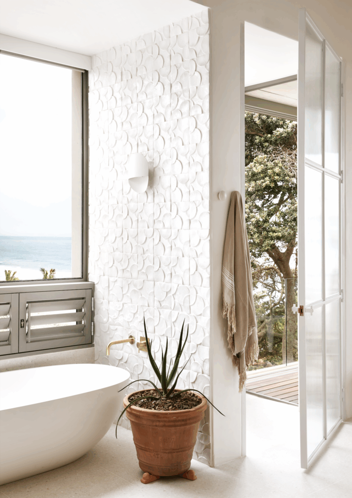 Handmade white Wheel of Time ceramic tile feature wall in a luxury bathroom with freestanding bathtub, textured artisan tiles and contemporary coastal styling.